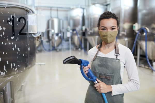 Waist up portrait of young female worker wearing mask and looking at camera while washing equipment at industrial factory, copy space